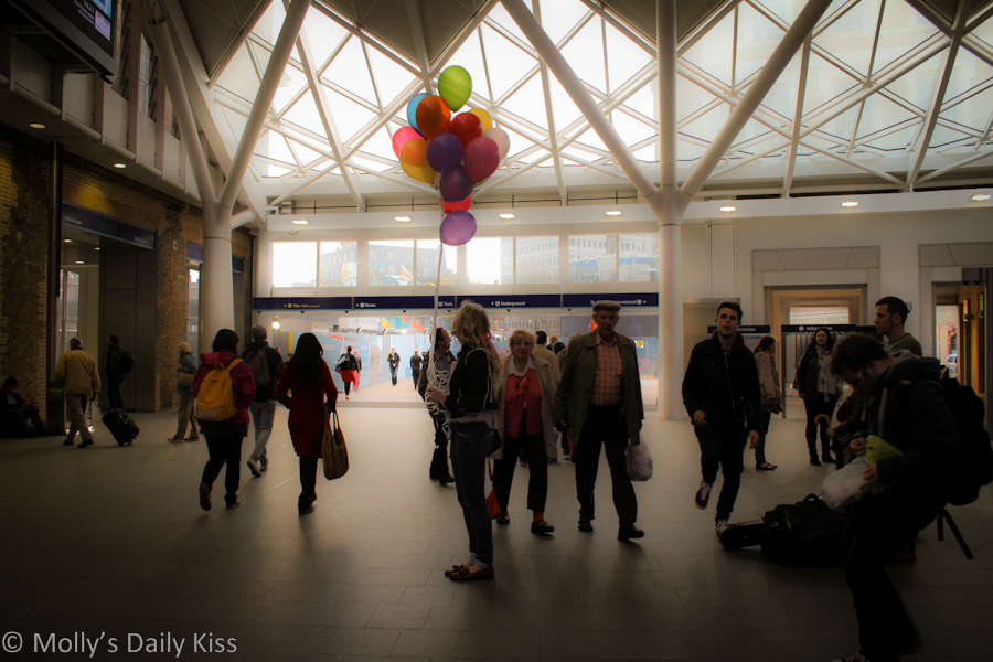 Woman at train station holding lots of coloured balloons